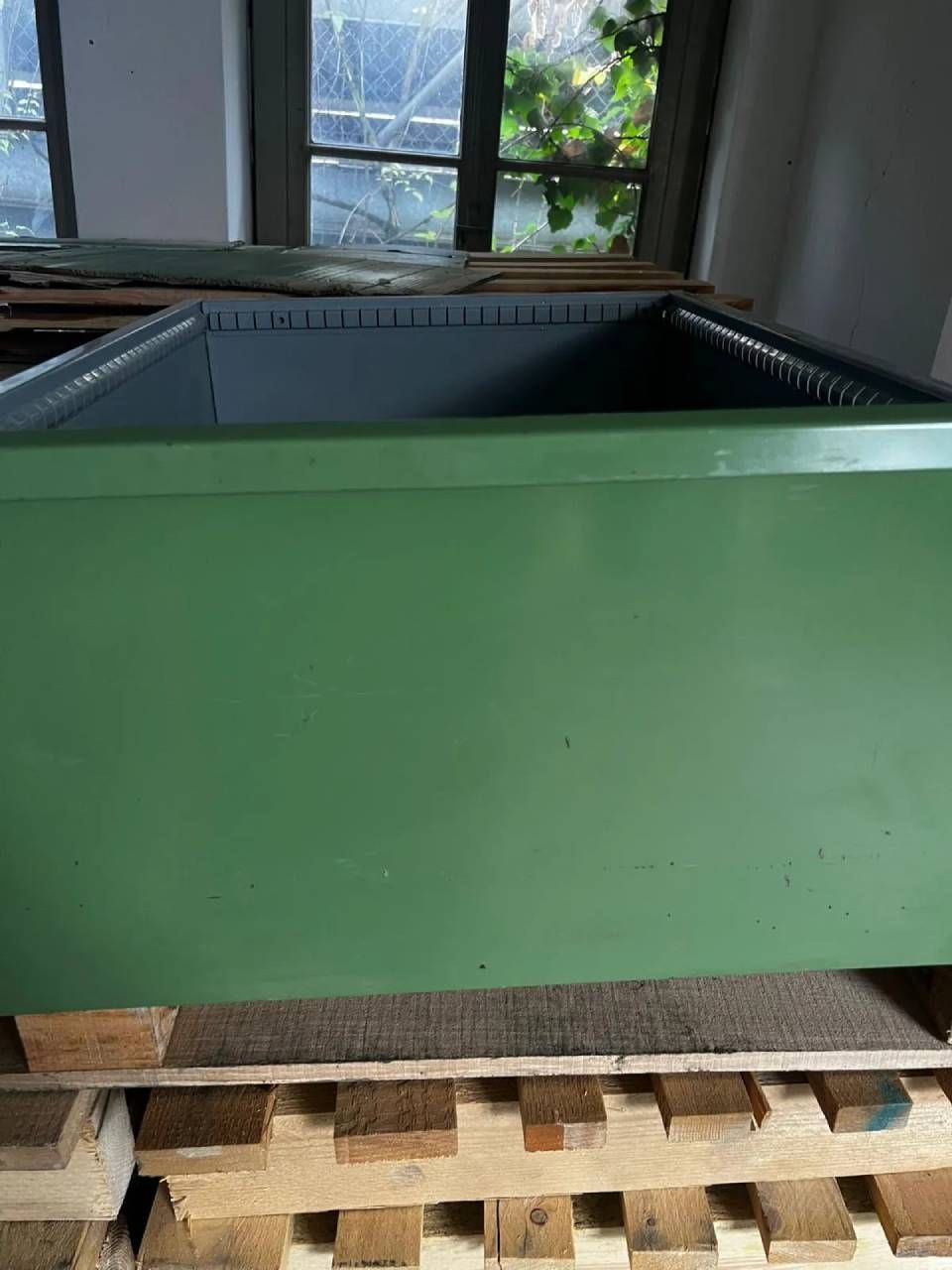 Ein grosser, oben offener grüner Metallbehälter, der auf einer Holzpalette in einer Werkstatt oder einem Industriebetrieb steht.