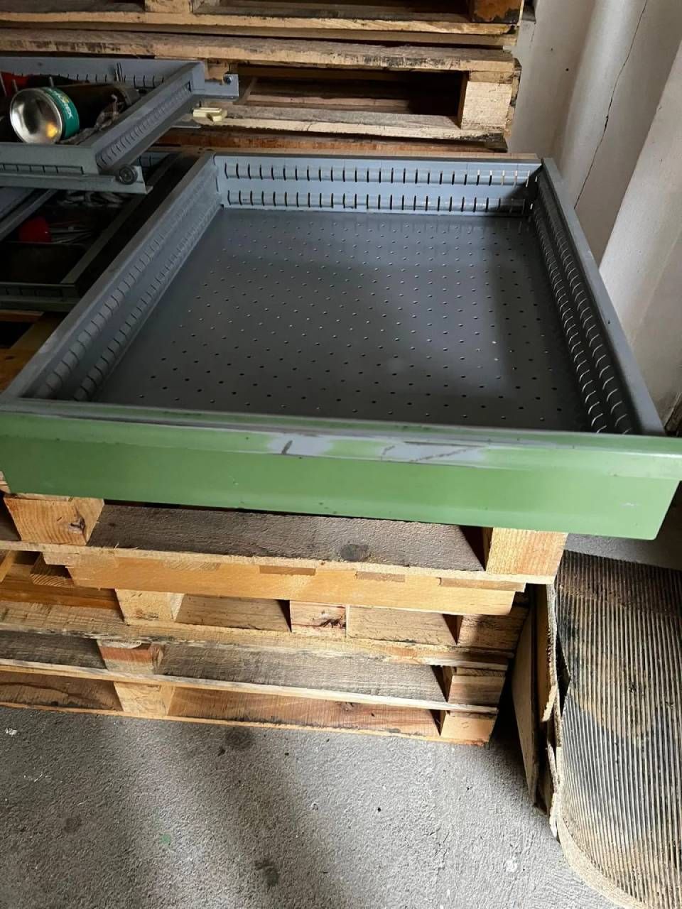 Ein graues Metalltablett mit perforierten Seiten und einer grünen Frontplatte, das auf gestapelten Holzpaletten ruht.