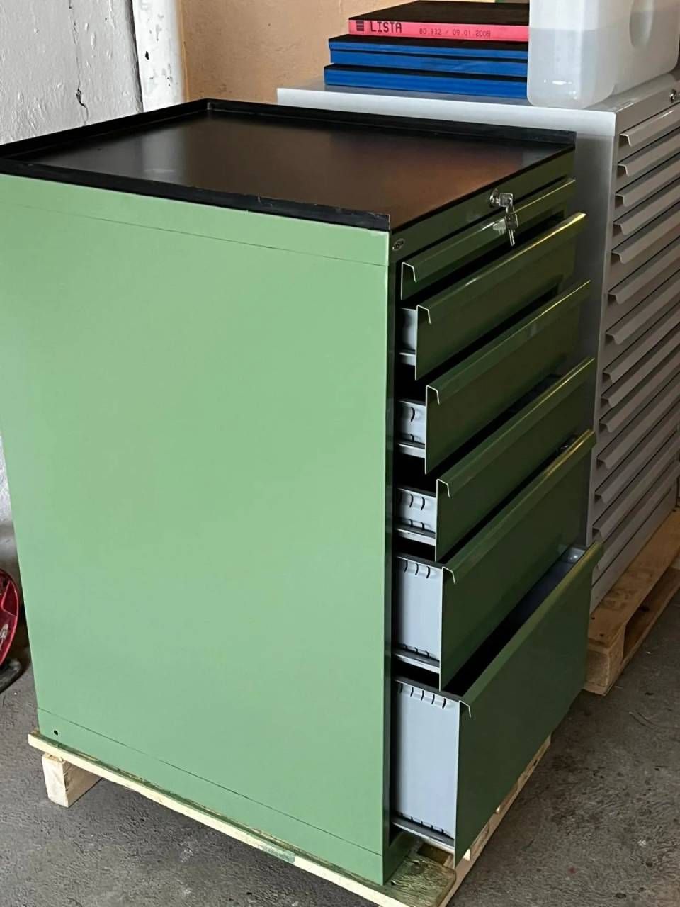 Ein hellgrüner Metallschrank mit fünf offenen Schubladen, der auf einer Holzpalette in einem industriellen Lagerraum steht.