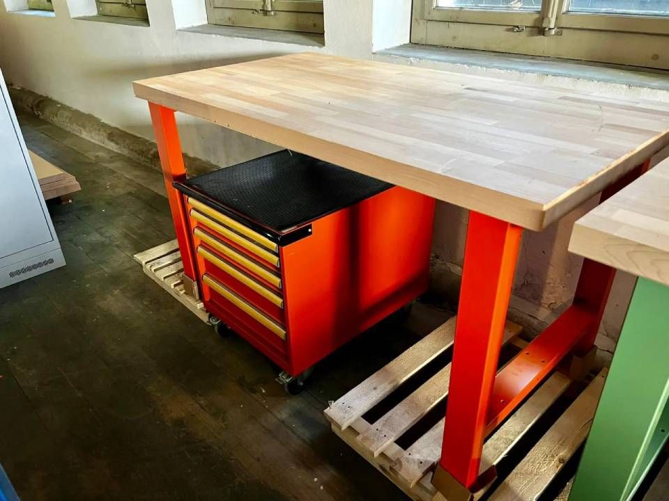 Auf einem Betonboden steht über einem fahrbaren roten Werkzeugschrank eine hölzerne Werkbank mit leuchtend orangefarbenen Beinen.