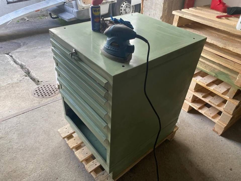 Ein hellgrüner Metallschrank steht auf einer Holzpalette, darauf ein handgeführter elektrischer Schleifer.