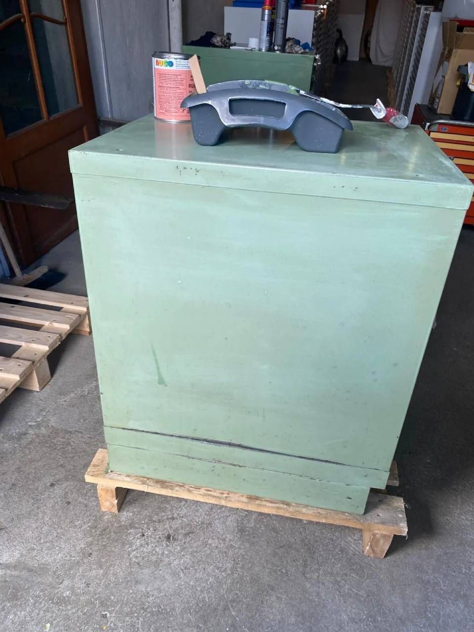 In einer Werkstatt steht auf einer Holzpalette ein salbeigrüner Metallschrank, darauf ein grauer Kartenleser und eine Farbdose.