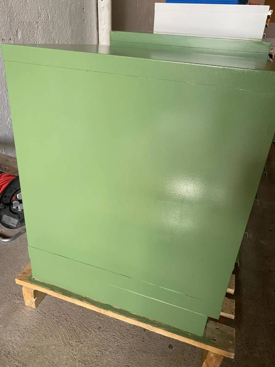 Ein mintgrüner Industrieschrank aus Metall steht auf einer hölzernen Versandpalette in einem Innenraum.