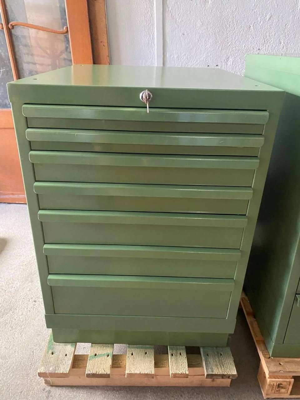 Ein hoher, olivgrüner industrieller Metallschrank mit mehreren Schubladen und einem zentralen Schloss, der auf einer Holzpalette steht.