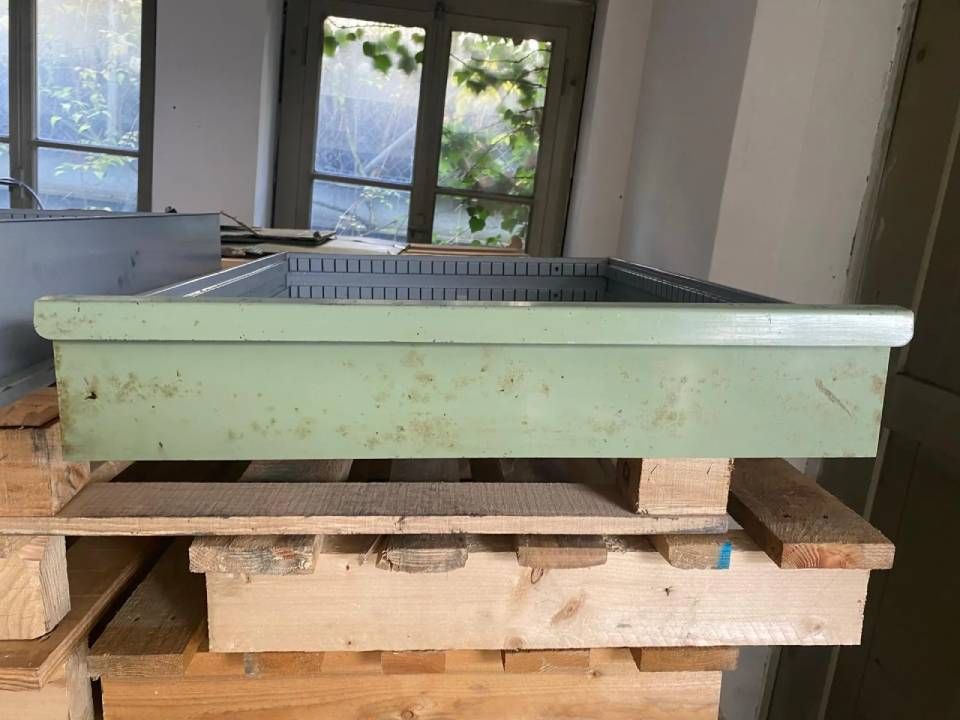 Ein hellgrünes, rechteckiges Metalltablett steht auf einer Holzpalette in einem Raum mit Fenstern im Hintergrund.