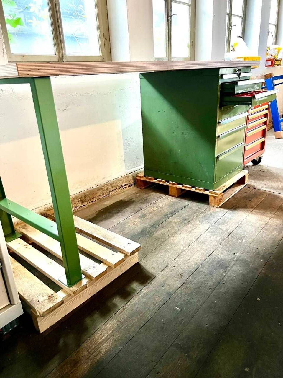 Eine hölzerne Werkbank mit grünen Metallbeinen und einem Schubladenelement, beide auf Holzpaletten in einem hellen, rustikalen Raum.
