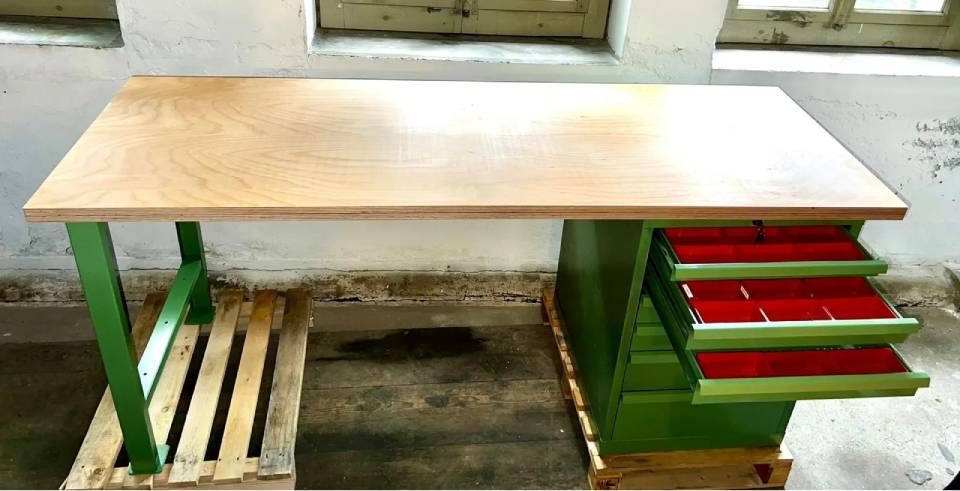 Eine Werkbank mit heller Holzplatte, grünen Beinen, einem Lattenrost aus Palettenholz auf der linken Seite und drei rot ausgekleideten Schubladen auf der rechten Seite.