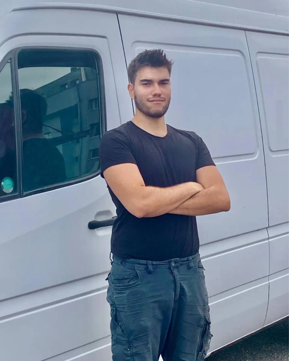 Eine Person mit verschränkten Armen steht lächelnd vor einem draussen geparkten weissen Lieferwagen.