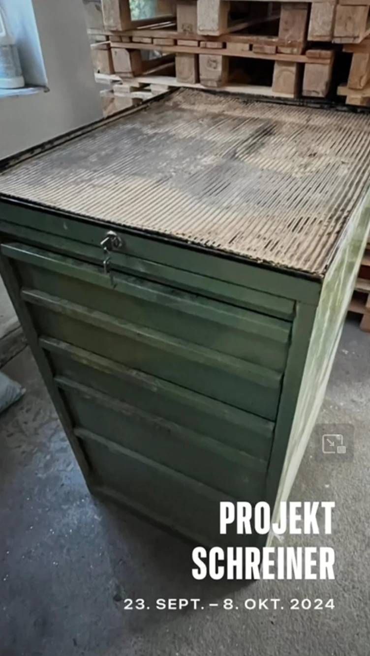 Ein olivgrüner Industrieschrank aus Metall mit mehreren Schubladen und einem abgenutzten Gitterrost obendrauf, in einer Werkstattumgebung.