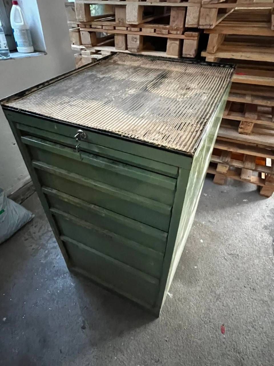 Ein grüner Vintage-Metallschrank im Industriestil mit fünf Schubladen und einer strukturierten Metallplatte, der in einem Abstellraum steht.