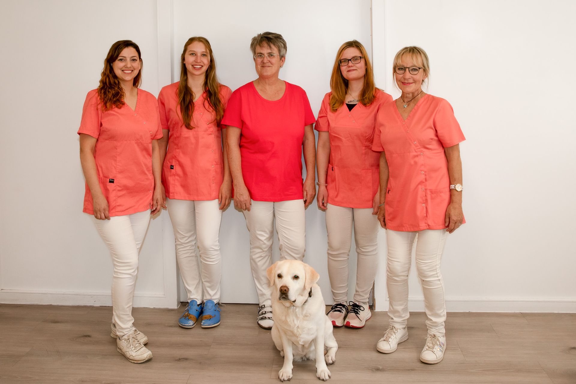 Fünf Frauen in rosa OP-Kleidung und weißen Hosen mit einem Golden Retriever vor einer weißen Wand.