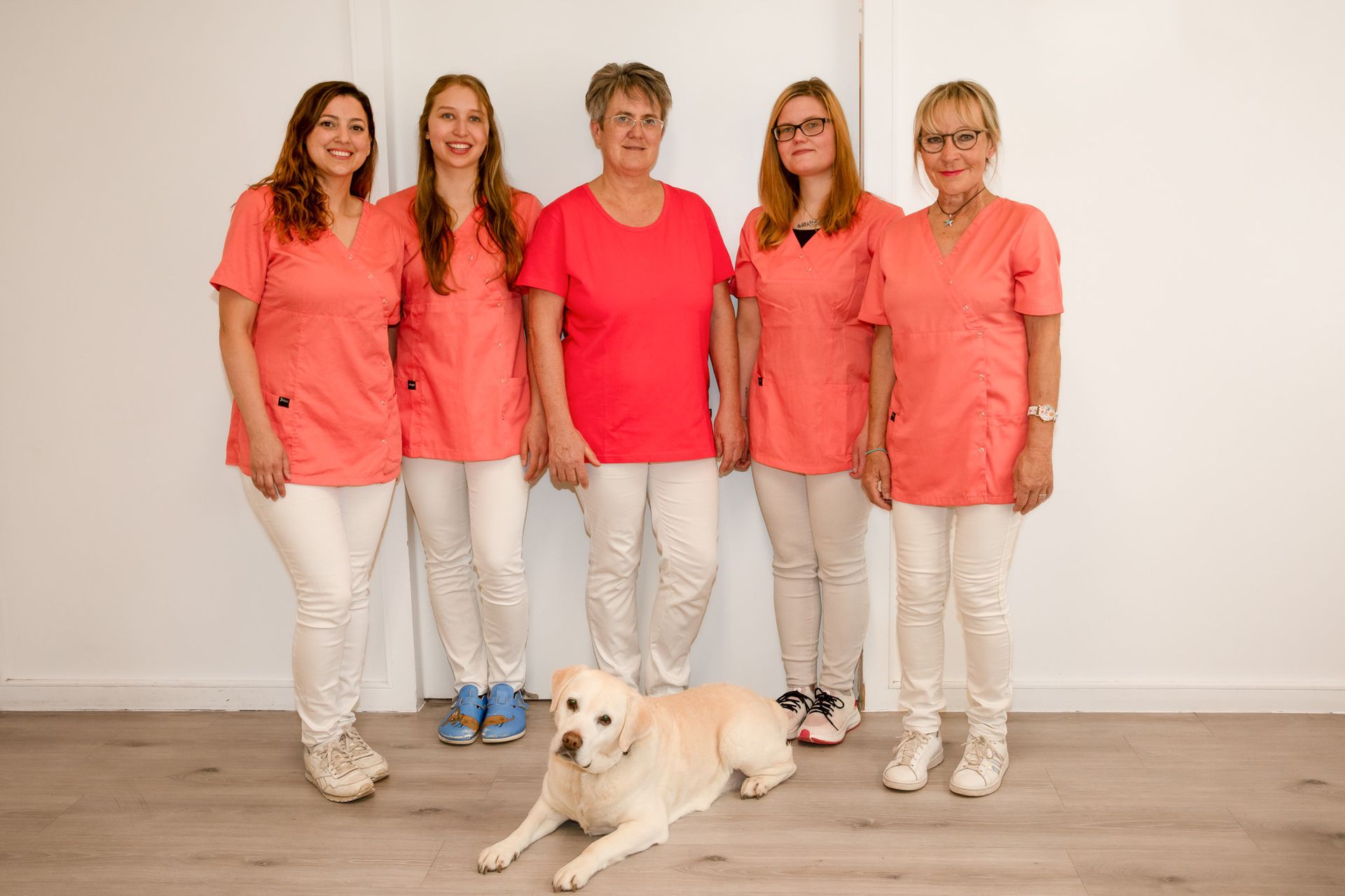 Fünf Frauen und ein Hund in passenden korallenfarbenen OP-Kleidern posieren vor einer weißen Wand.