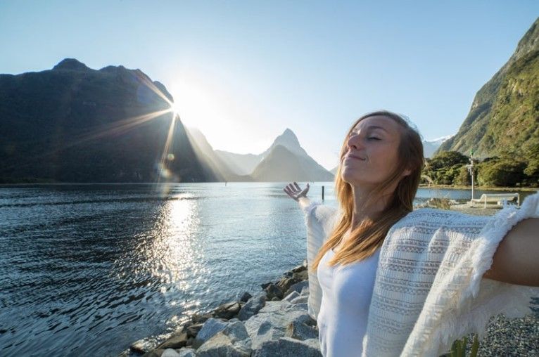 Mujer con los brazos extendidos disfrutando del sol cerca de un lago y montañas.