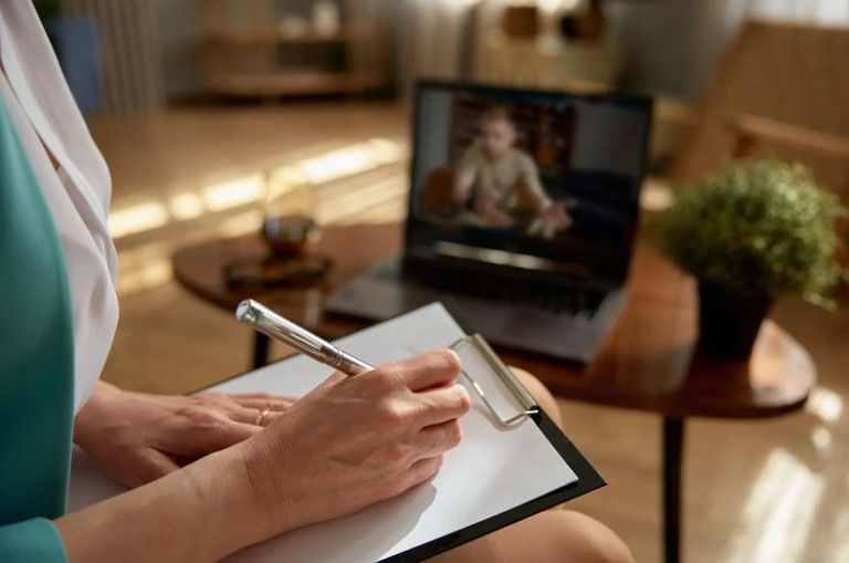 Persona tomando notas durante una sesión virtual con un niño en un ordenador portátil en un salón bien iluminado.