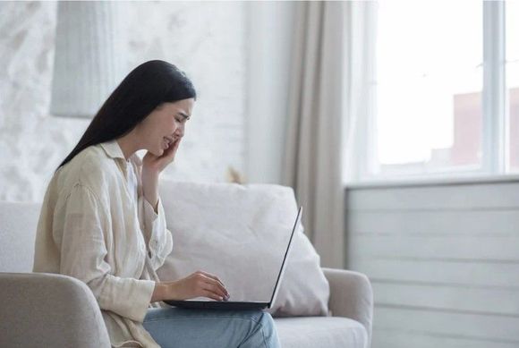 Mujer sentada en un sofá, usando una computadora portátil, tocándose la cara. Habitación luminosa de colores neutros con una ventana.