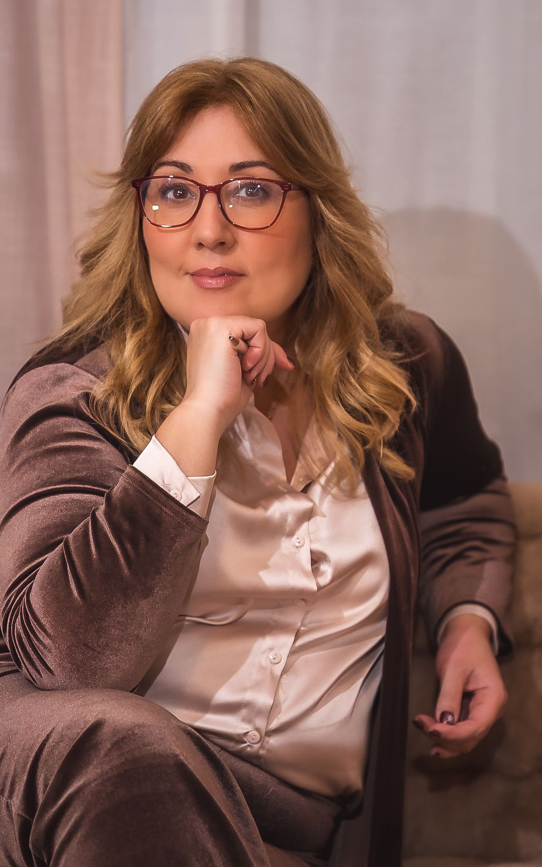 Mujer con gafas sonriendo a la cámara, sentada cerca de un sofá en una habitación luminosa.