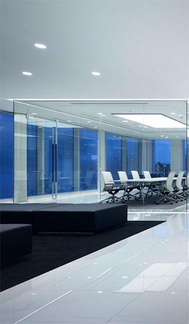 Interior de oficina moderno con paredes de cristal, mesa de conferencias y asientos negros. Exterior azul visible a través de las ventanas, techo y suelo blancos.