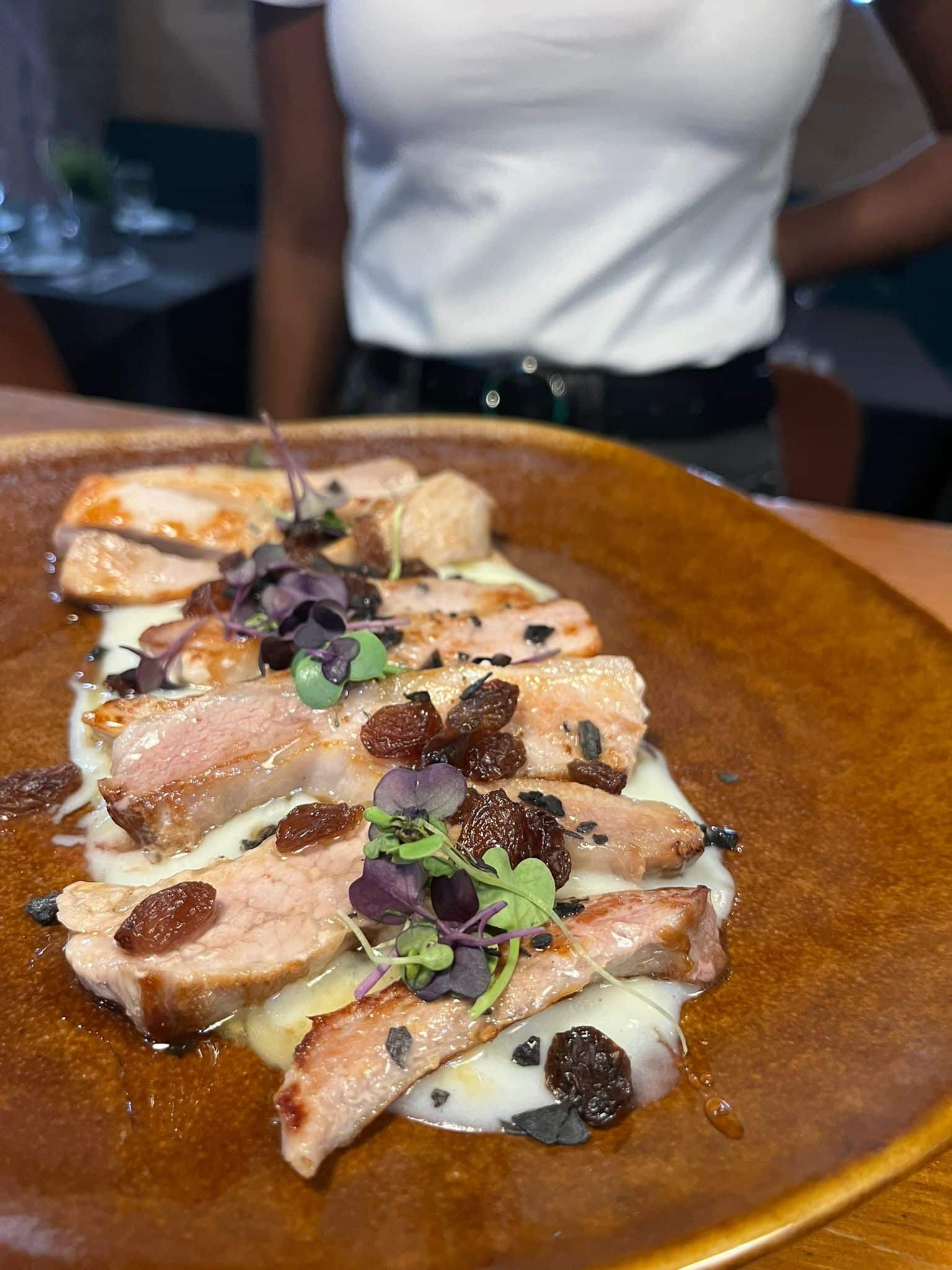 Cerdo en rodajas con salsa, cubierto con bayas y hierbas, en un plato marrón, persona en el fondo.