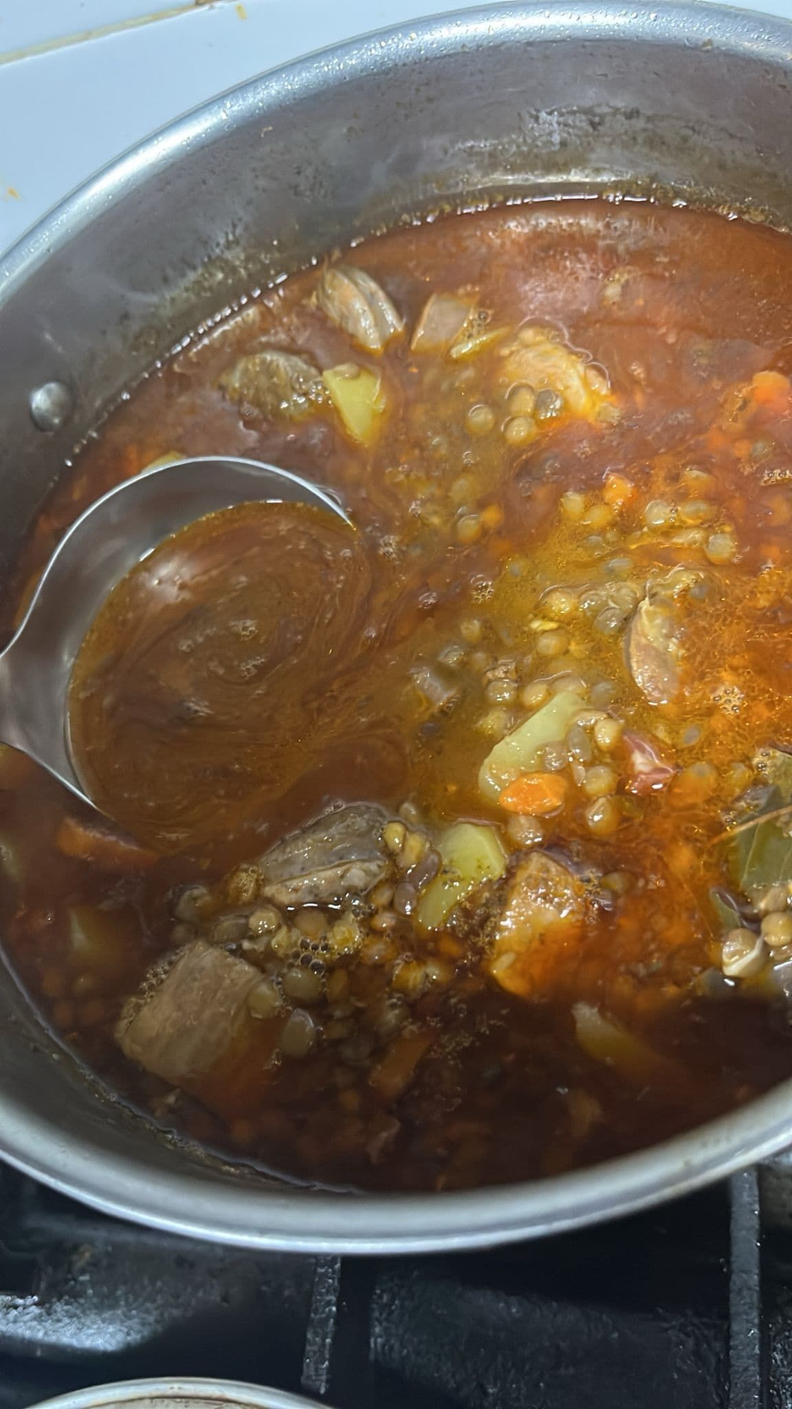 Una olla de sopa de lentejas con carne servida con un cucharón. El guiso tiene trozos visibles de carne, papas y zanahorias.