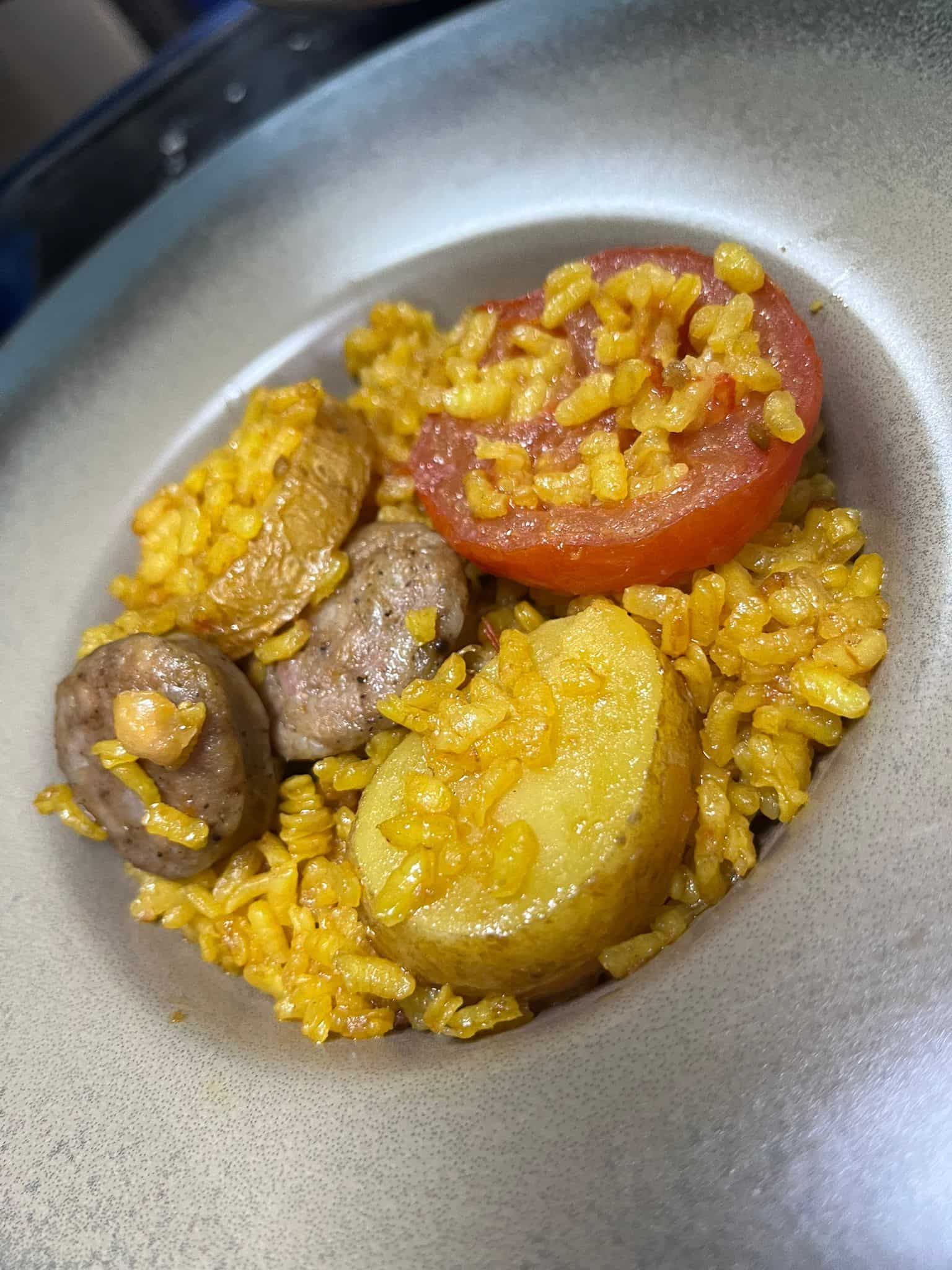 Plato de arroz amarillo, patatas, salchicha y medio tomate en un recipiente gris.