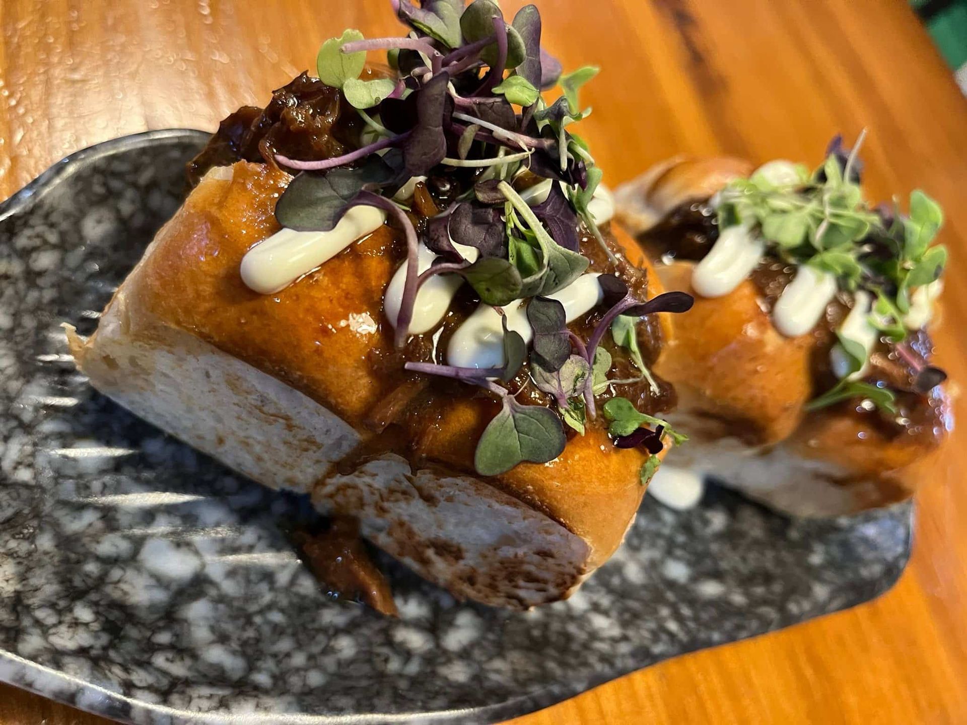 Dos hamburguesas con salsa oscura, microvegetales y llovizna blanca sobre un plato gris estampado.