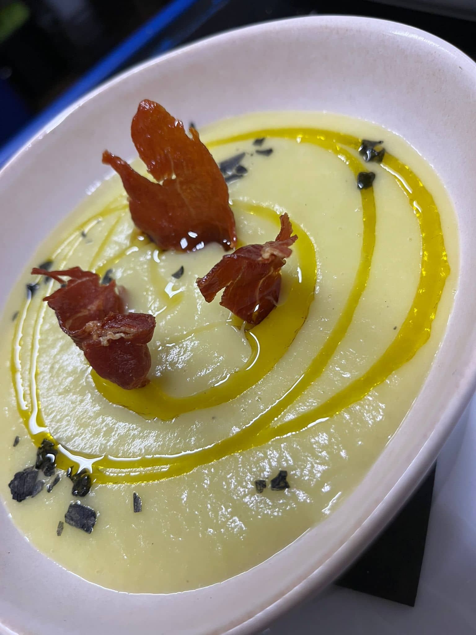 Sopa cremosa con jamón y un chorrito de aceite de oliva, adornada con hojuelas negras en un tazón blanco.