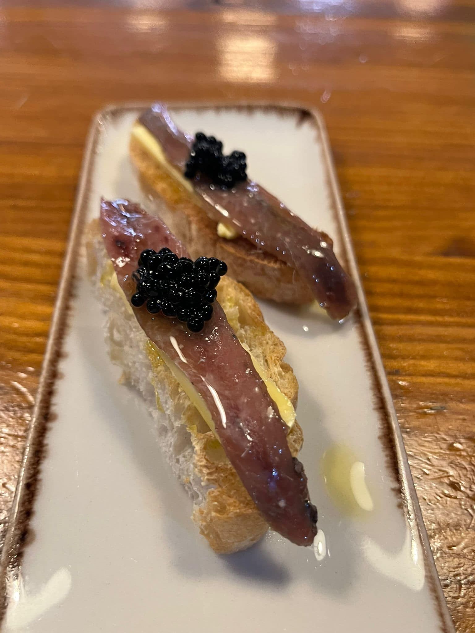 Dos tapas en un plato rectangular blanco: pan cubierto con anchoa, caviar y mantequilla.