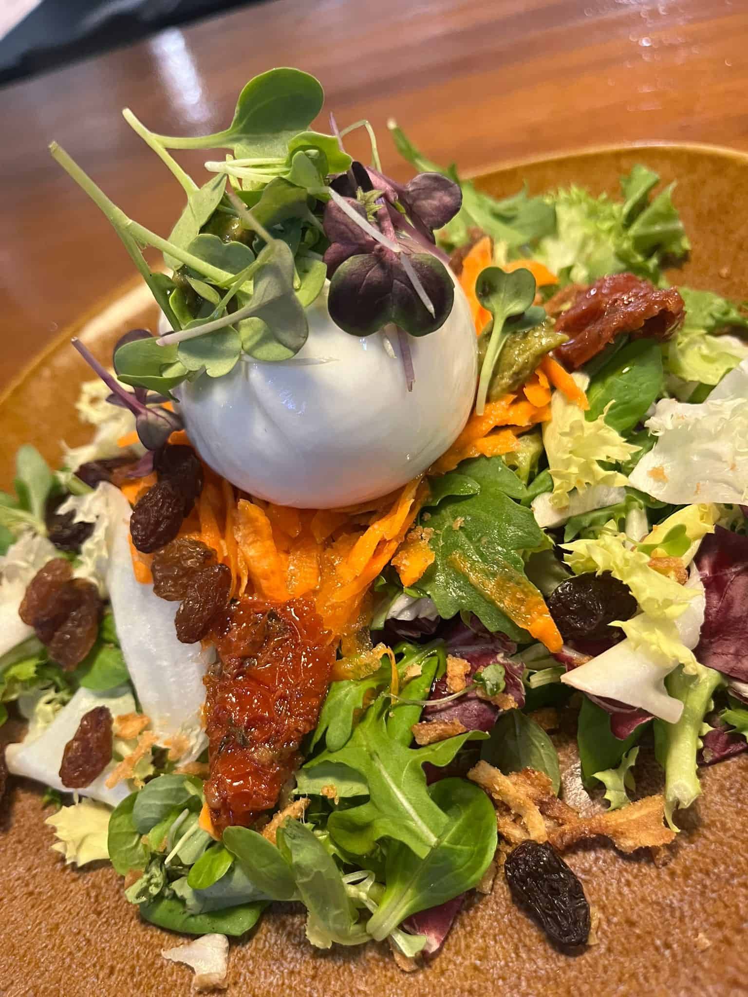 Ensalada con huevo escalfado, tomates secados al sol, zanahorias ralladas, pasas y verduras en un plato marrón.