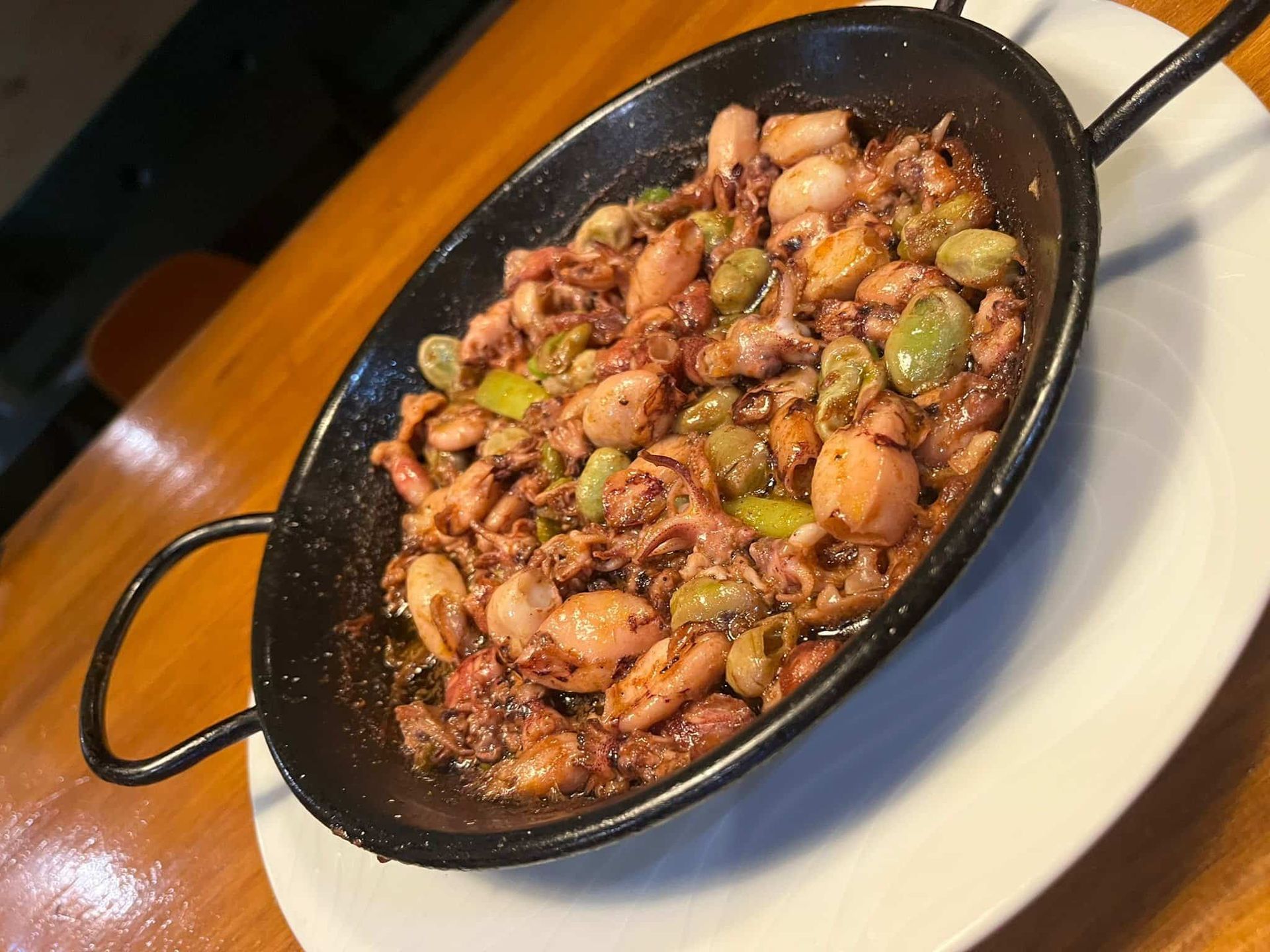 Plato de calamares y habas en una sartén negra sobre un plato blanco, servido en una mesa de madera.