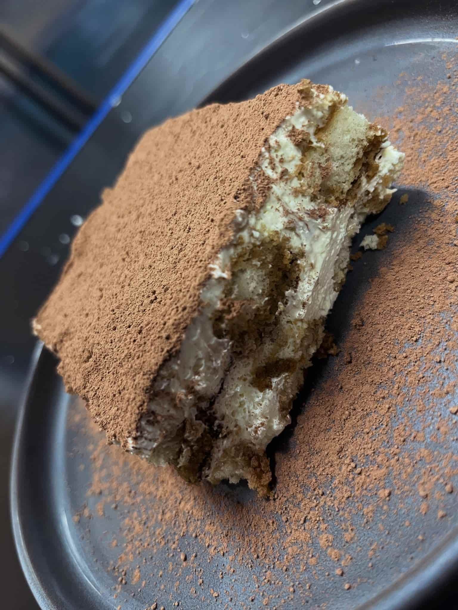 Rebanada de tiramisú en un plato oscuro, espolvoreada con cacao en polvo, mostrando capas de pastel y crema.