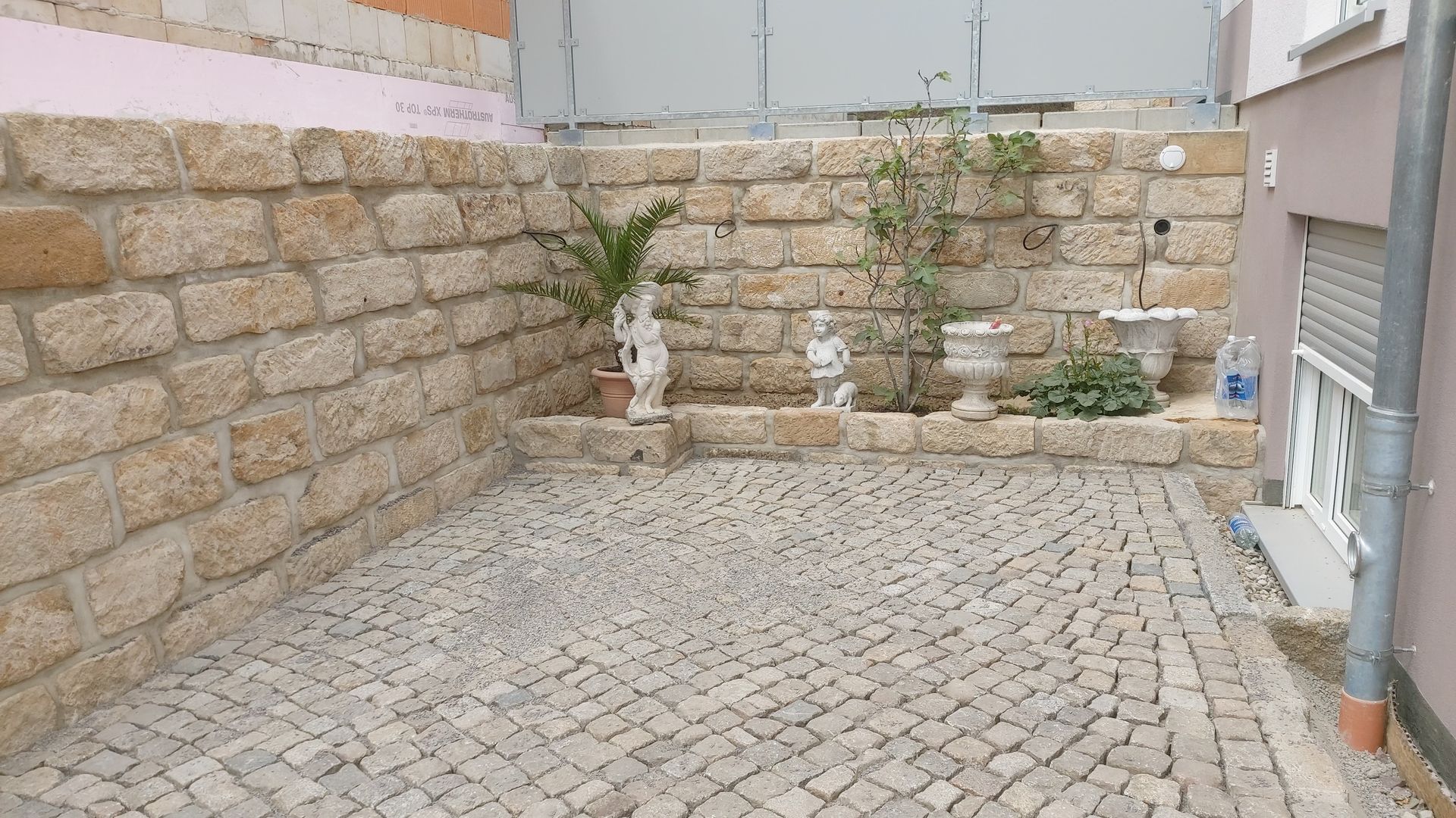 Gepflasterter Hof mit Steinstützmauer, Pflanzen und dekorativen Statuen.