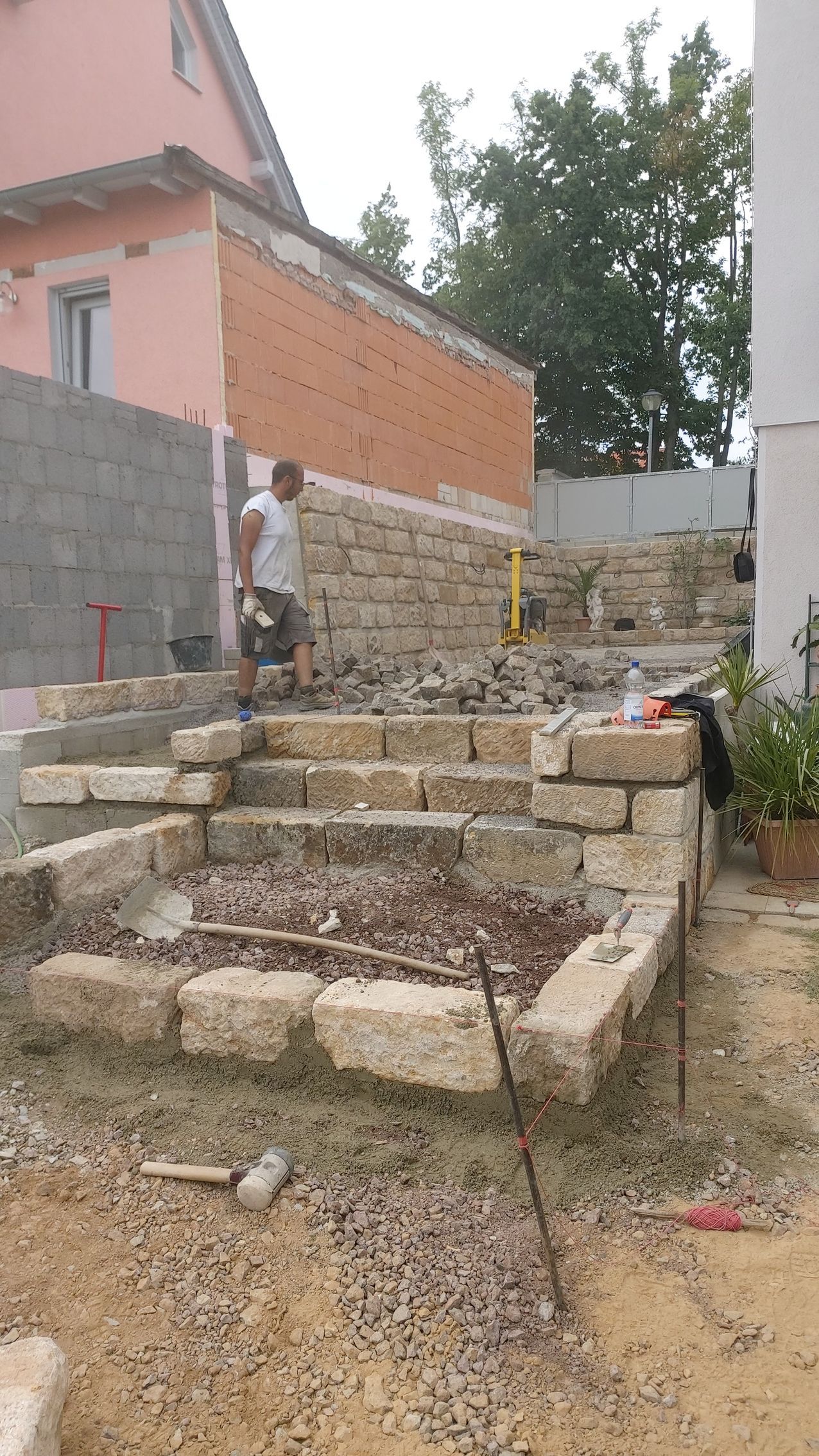 Mann baut Steinstufen in einem Hof, mit Stützmauer und rotem Backsteingebäude im Hintergrund.