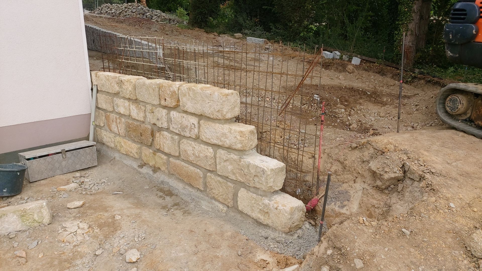 Neben einem Gebäude wird eine Stützmauer aus Stein gebaut. Erdarbeiten und Maschinen sind sichtbar.