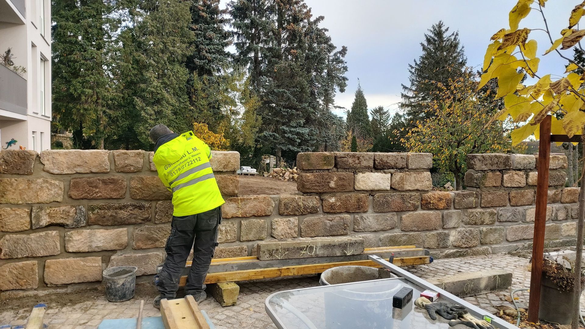 Ein Arbeiter in einer Warnweste baut im Freien neben einem Gebäude und Bäumen eine Steinmauer.
