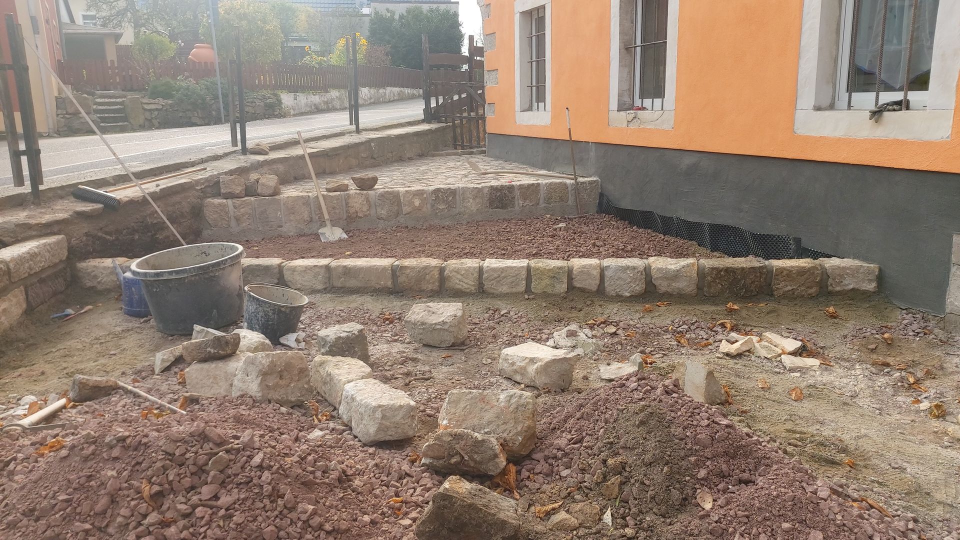 Baustelle mit Steinen, Erde und einem Gebäude. Eine Person scheint eine niedrige Mauer zu bauen.