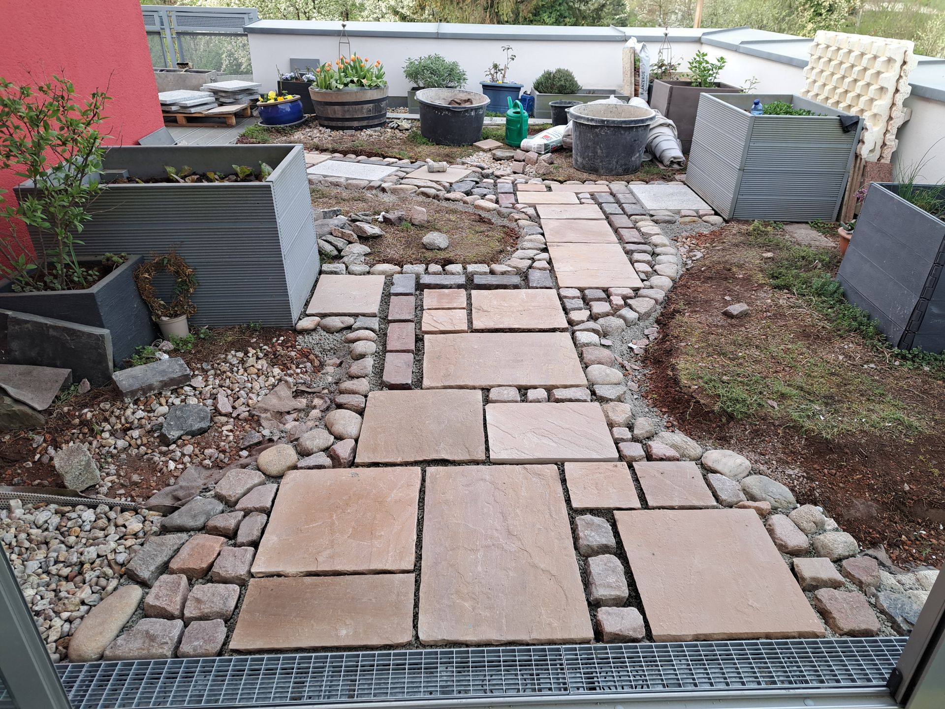 Steinweg durch einen kleinen Garten mit Hochbeeten und verschiedenen Pflanzen in Behältern.