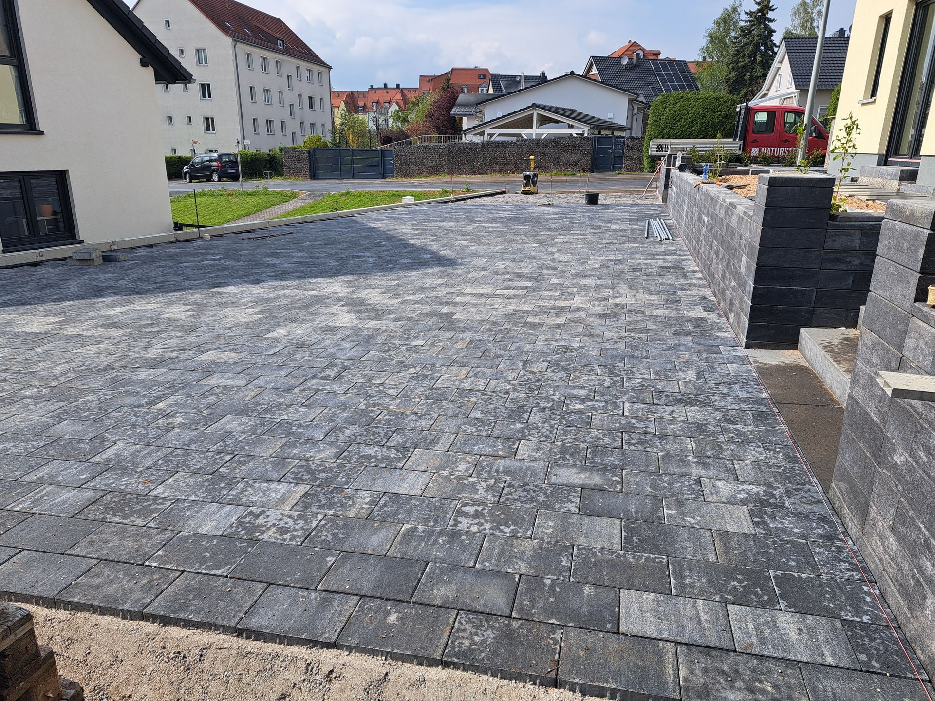 Neu gepflasterte Auffahrt mit dunkelgrauen Pflastersteinen, begrenzt durch eine Mauer, mit Gebäuden im Hintergrund.