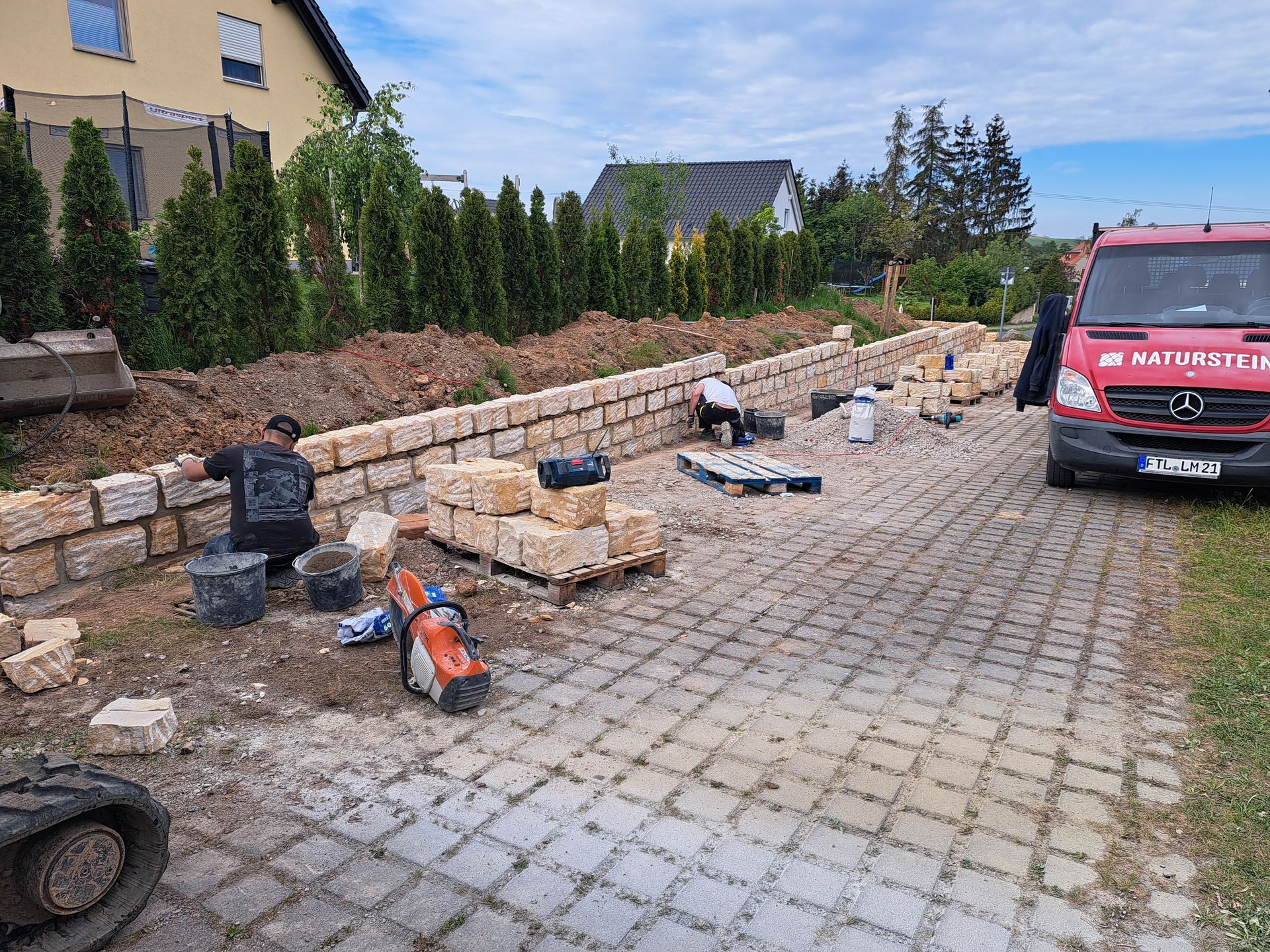 Bauarbeiter errichten eine Stützmauer aus Stein auf einer gepflasterten Auffahrt; in der Nähe parkt ein roter Lieferwagen.