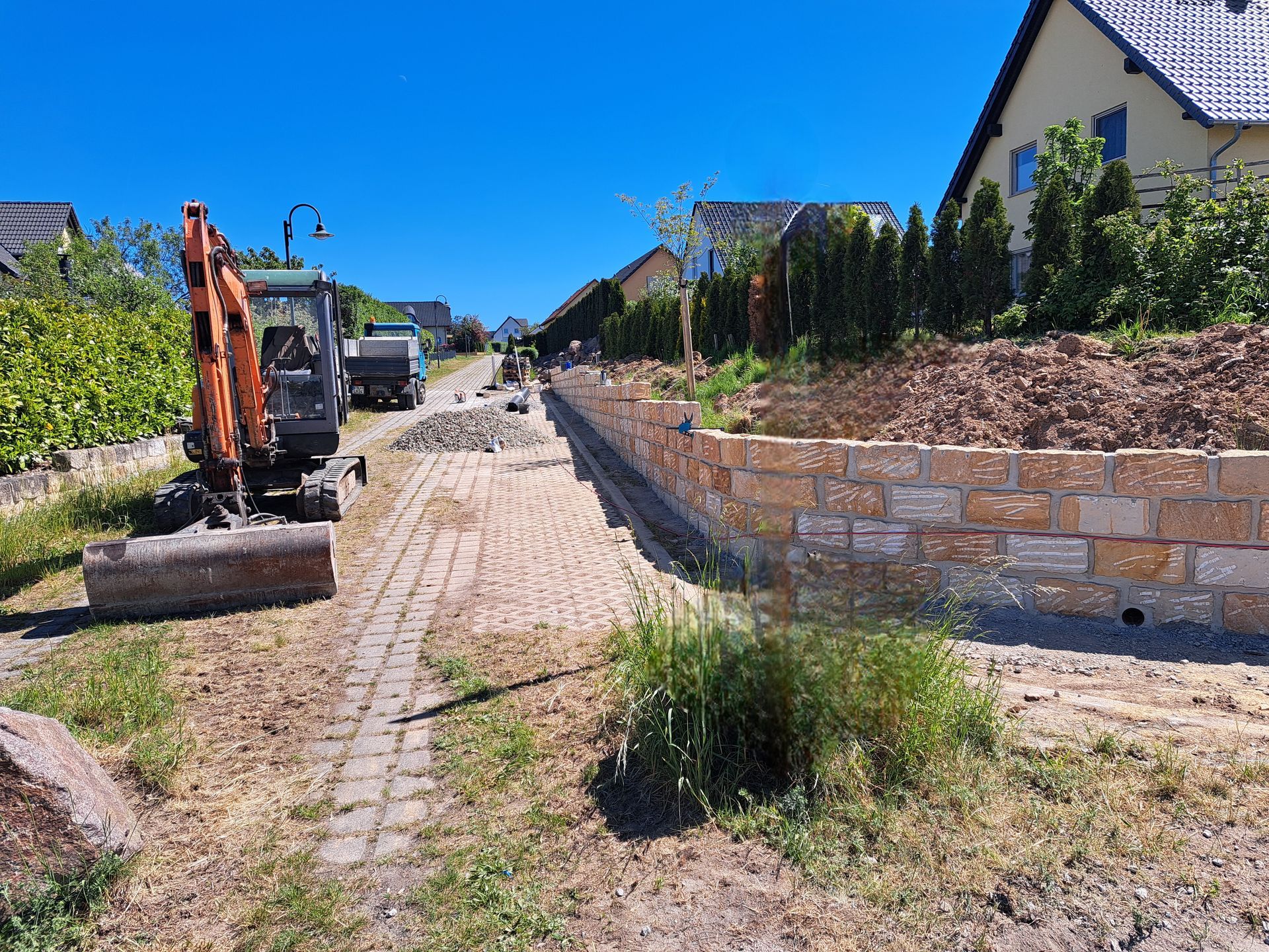 Baustelle: Minibagger auf Ziegelweg, Steinstützmauer, Wohnstraße unter blauem Himmel.