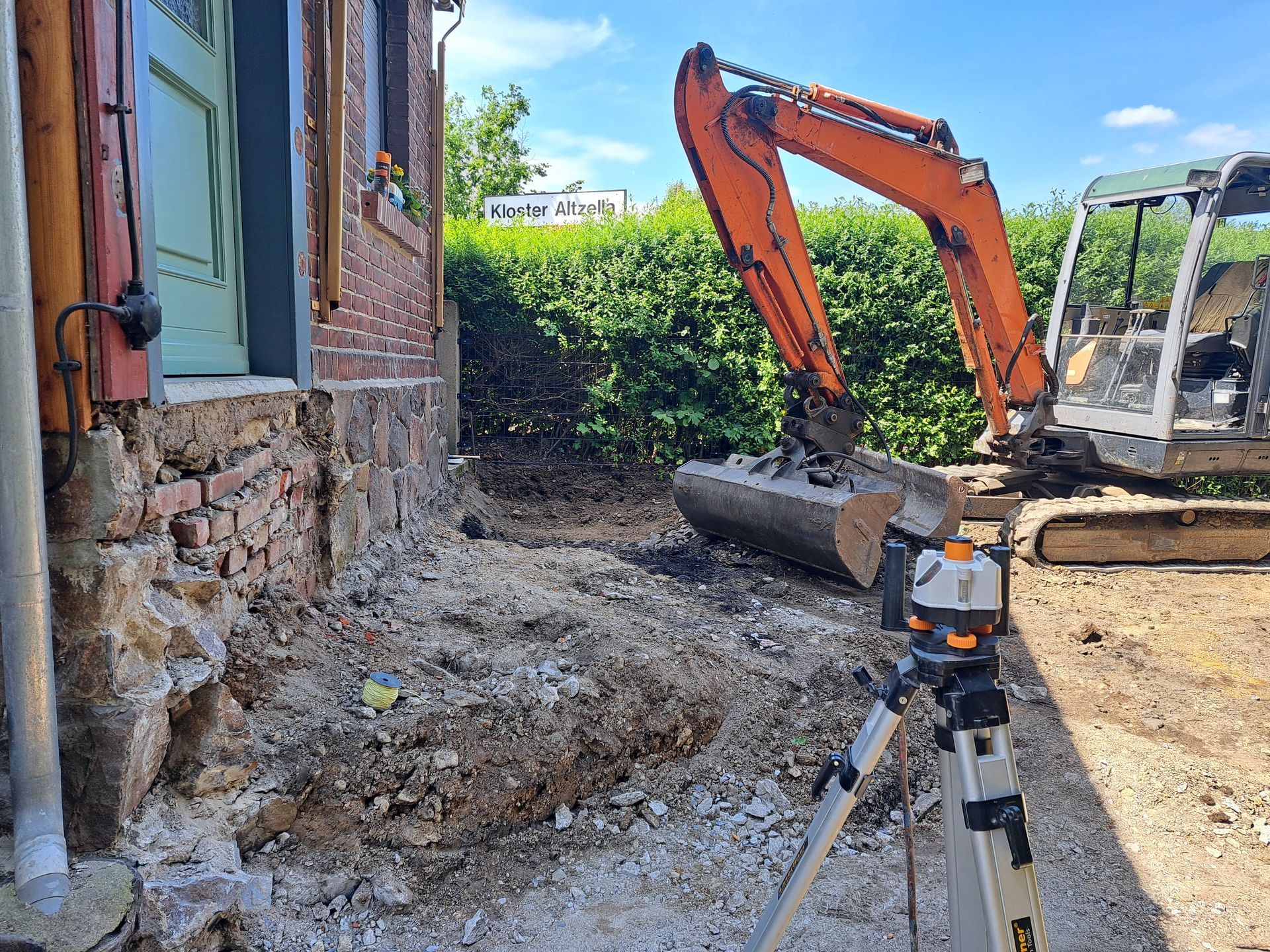 Bagger neben Backsteingebäude, der Erde ausgräbt. Im Vordergrund steht eine Wasserwaage. Sonniger Tag.