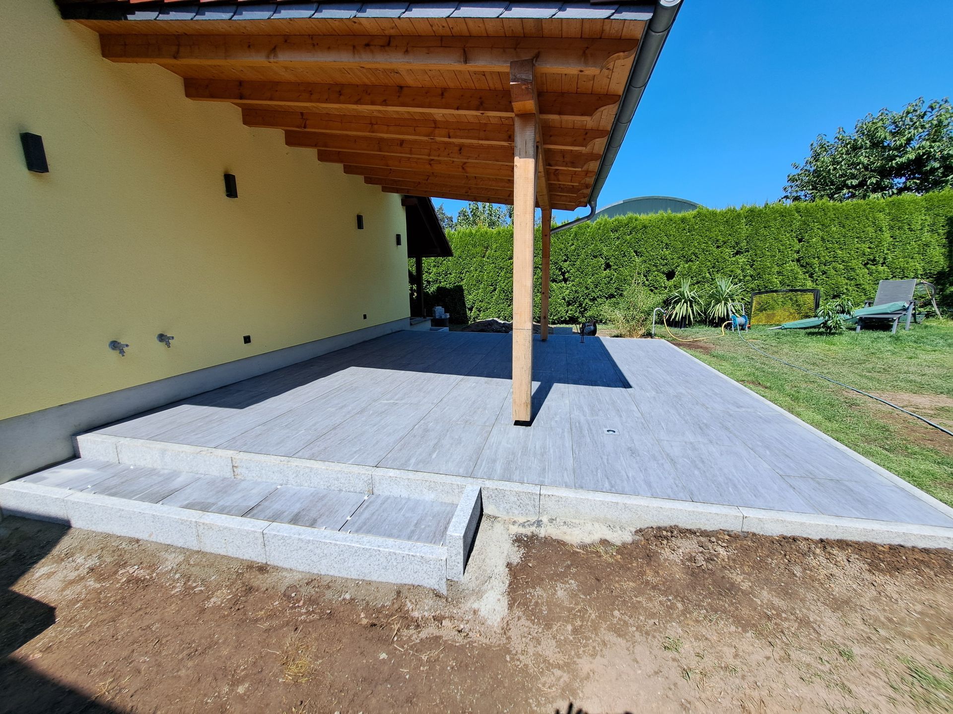 Holzterrasse mit Stufen neben einem gelben Haus, unter einem Holzbalkendach, mit einem grünen Hof.