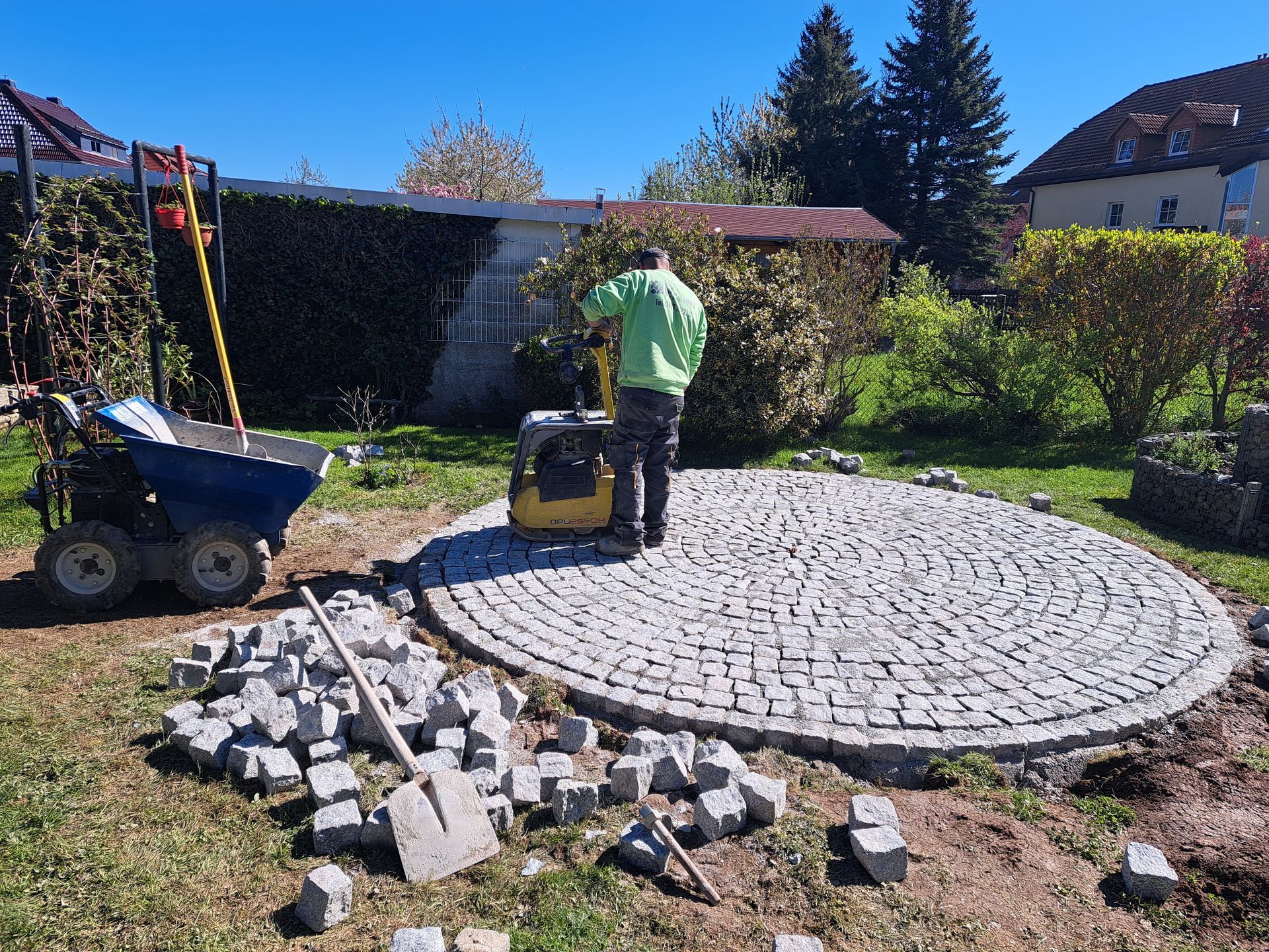 Mann verdichtet Kopfsteinpflaster in einem grasbewachsenen Hinterhof. 
