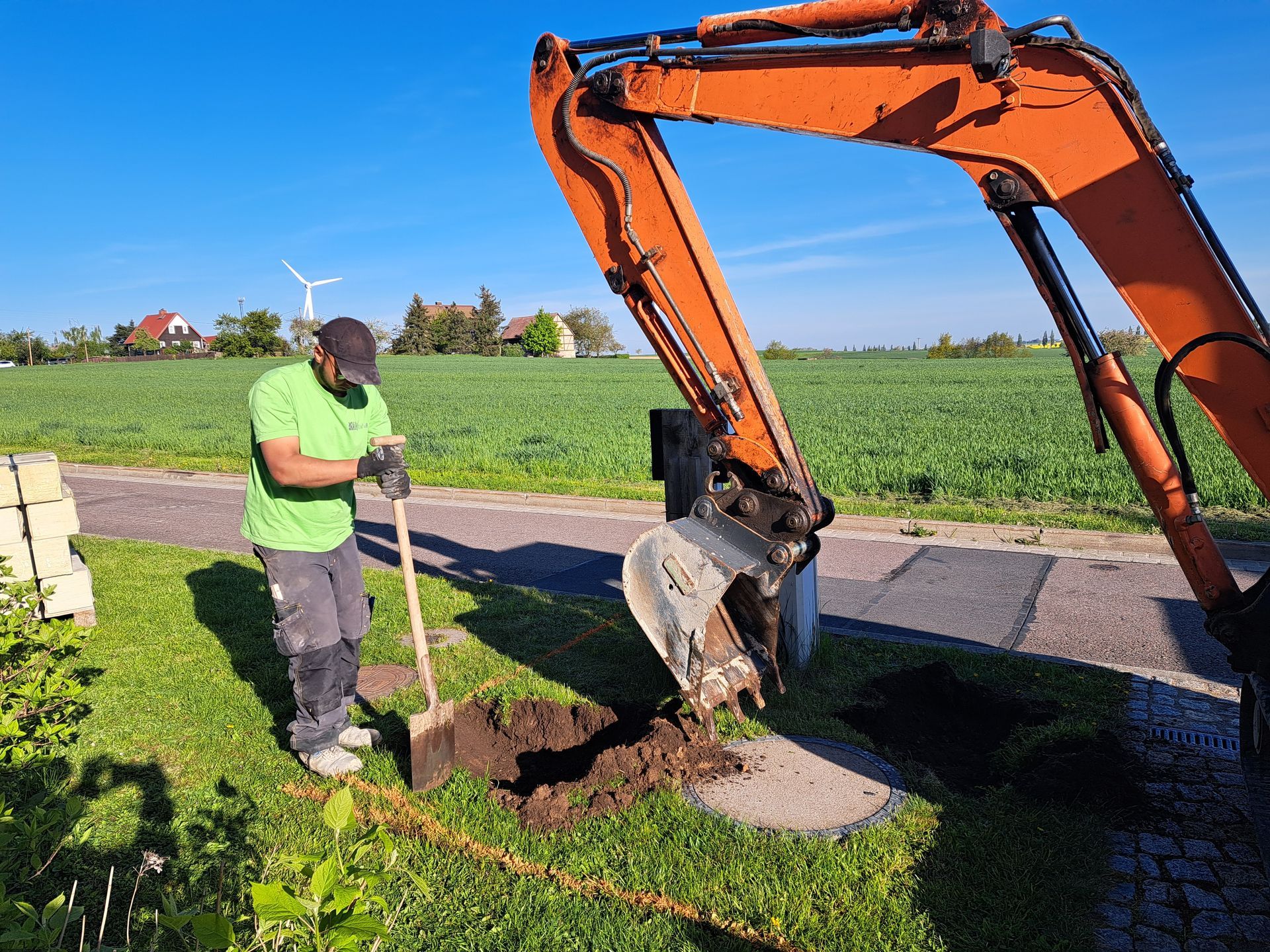 Mann gräbt mit Schaufel, Bagger in der Nähe; grünes Hemd, sonniger Tag, ländliche Umgebung.