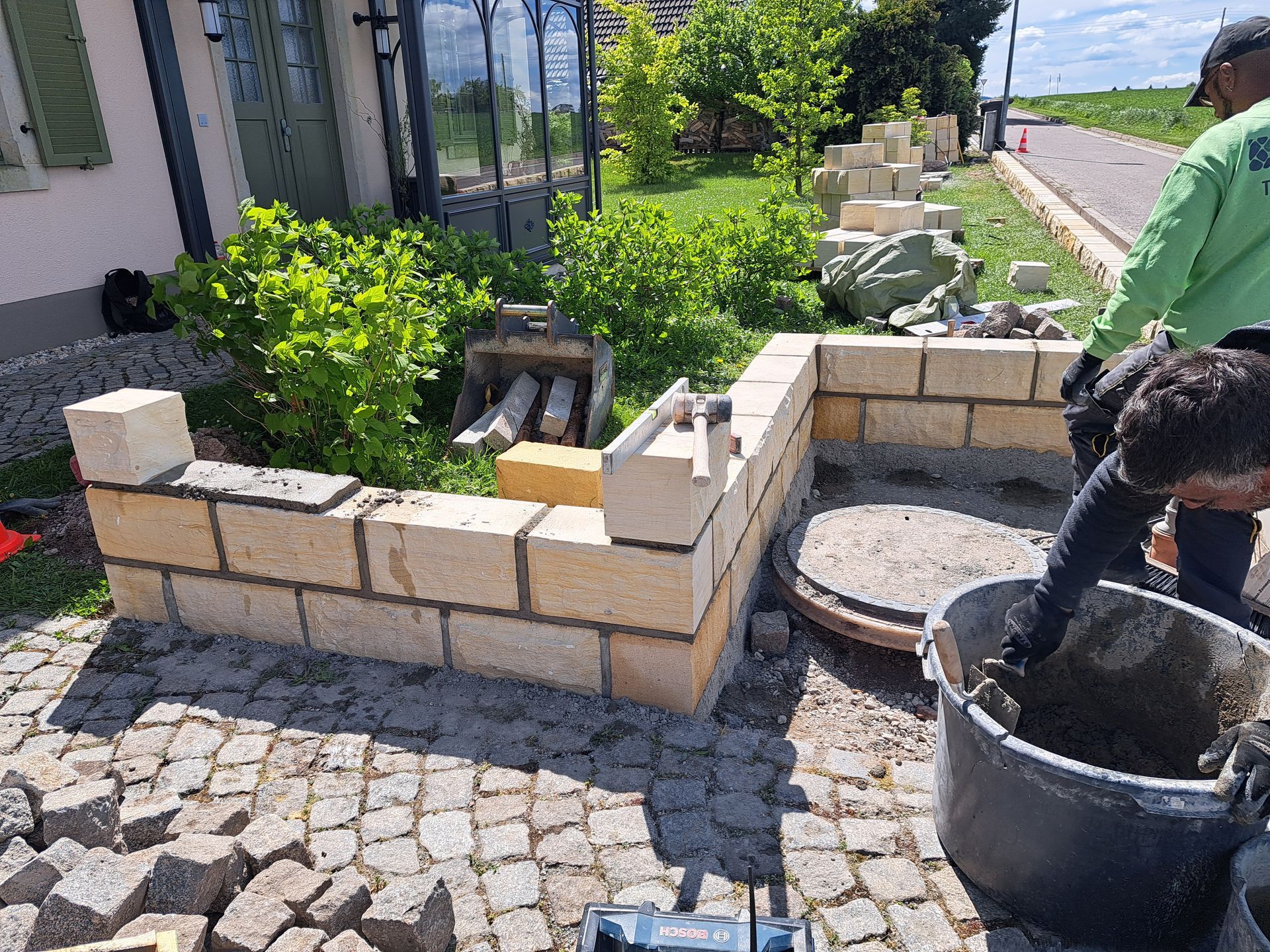 Bau einer Backsteinmauer um einen Garten. Eine Person mischt Mörtel in einem Eimer. Draußen, sonniger Tag.