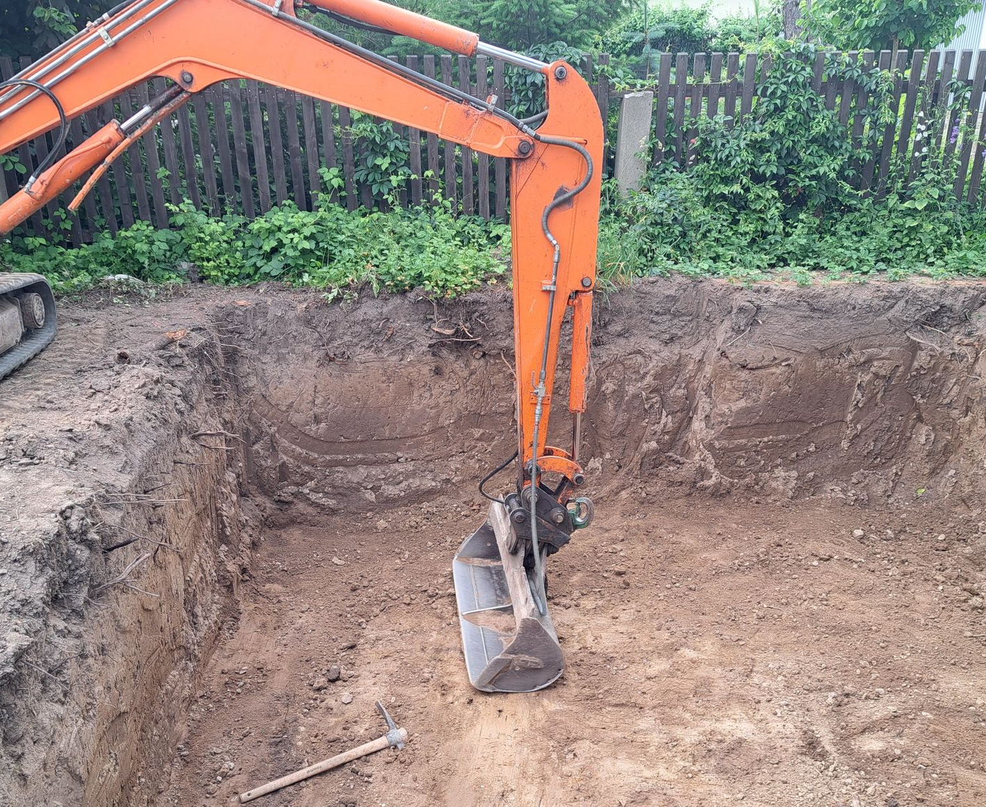 Eine orangefarbene Baggerschaufel in einem frisch gegrabenen quadratischen Loch. 