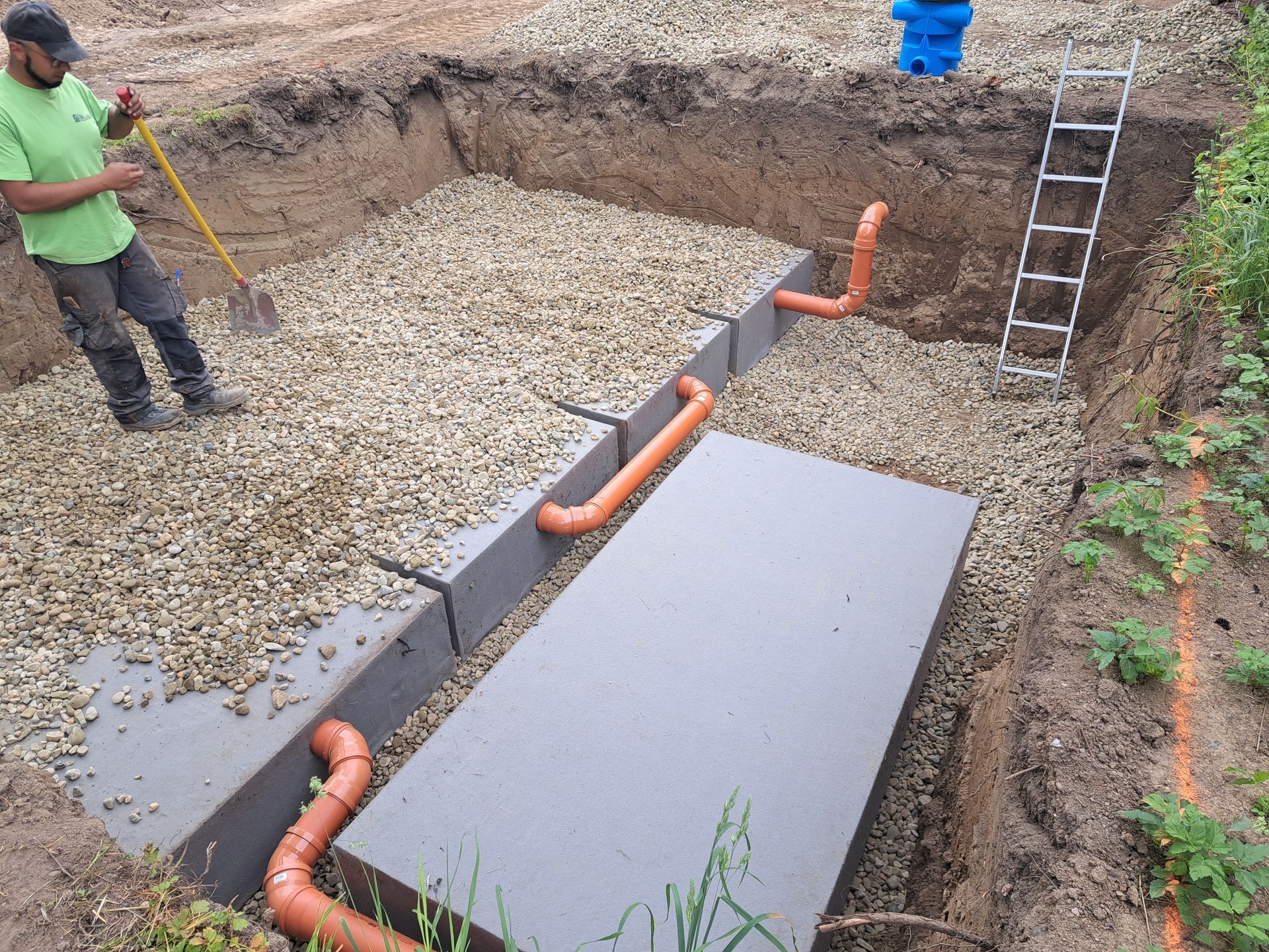 Mann schaufelt Kies in einen tiefen Graben; orangefarbene Rohre und graue Blöcke werden installiert.