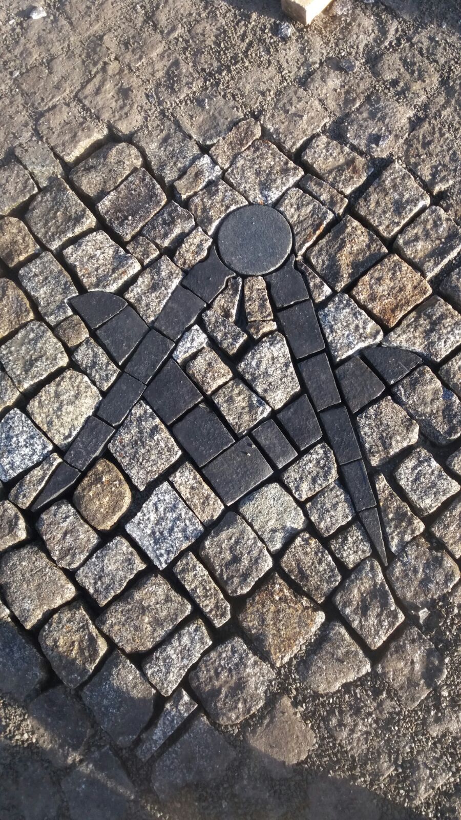 Freimaurersymbol, ein Zirkel und ein Winkel, gepflastert mit schwarzen Steinen vor einem Kopfsteinpflasterhintergrund.