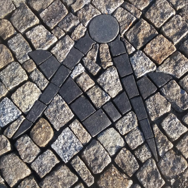 Winkel- und Zirkelsymbol aus dunklen Steinen, eingelegt in einen gepflasterten Gehweg.