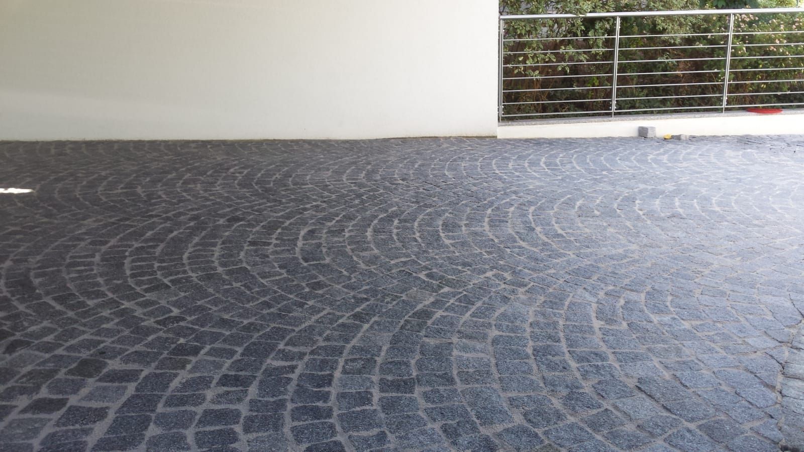 Dunkelgraues Kopfsteinpflaster mit Bogenmustern, neben einer weißen Wand und einem Balkon.