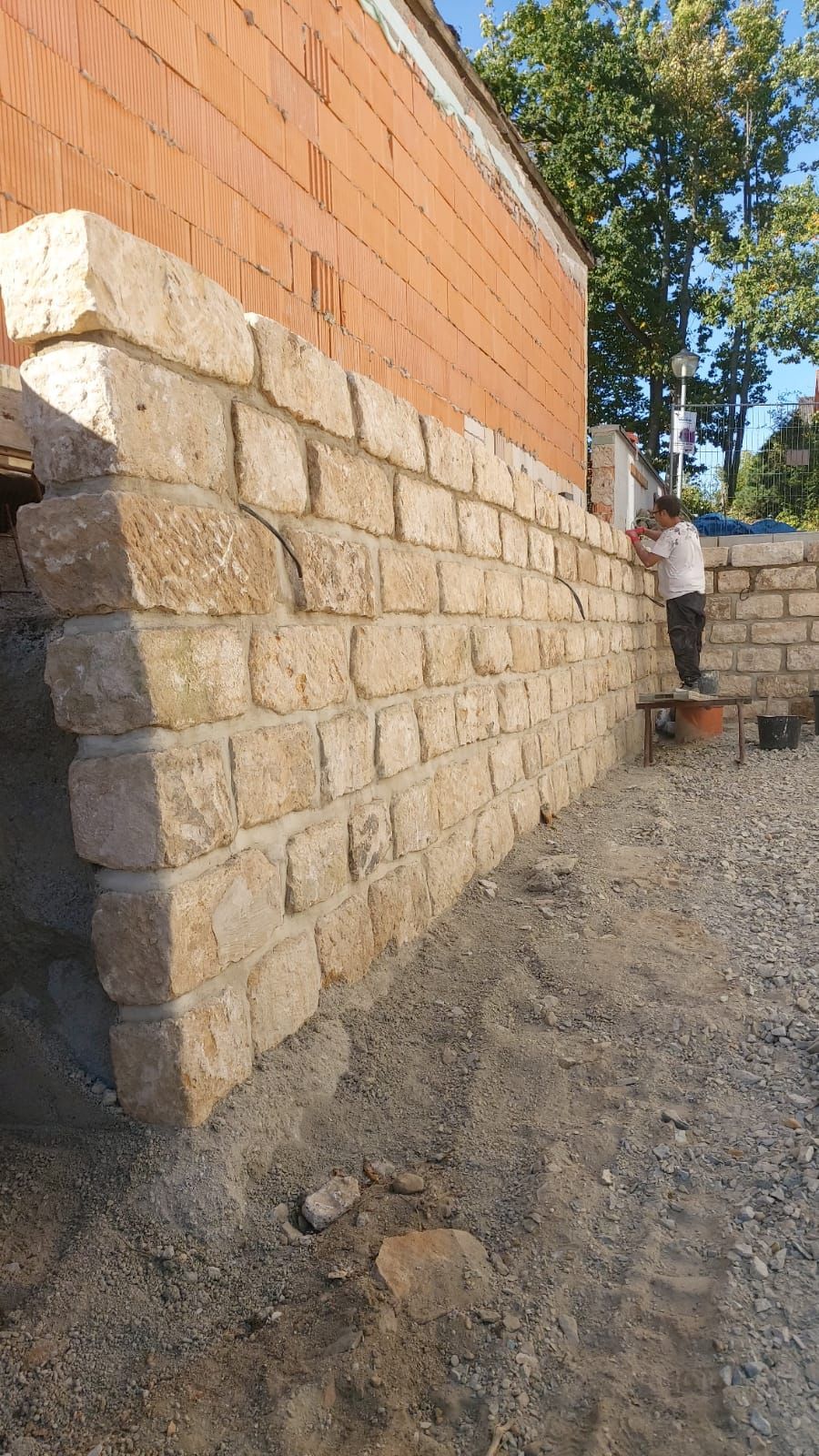 Neben einem Backsteingebäude wird eine Stützmauer aus Stein errichtet; ein Mann arbeitet an der Mauer.